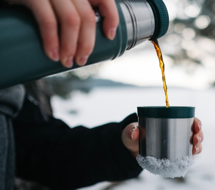 Hands holding an insulated thermos flask pouring tea or coffee into a rubber insulated metal cup against a snow background 