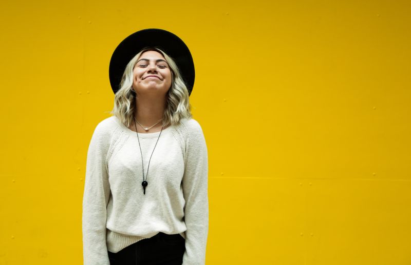 Yellow background with young woman with black rimmed hat, shoulder-length blonde wavy hair looking up and smiling