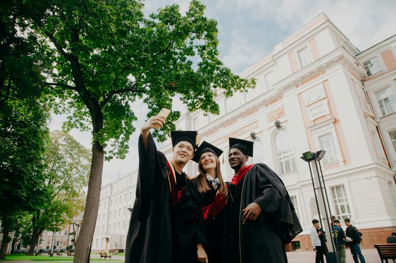 image of three students wearing caps and gowns and taking a selfie outside a university