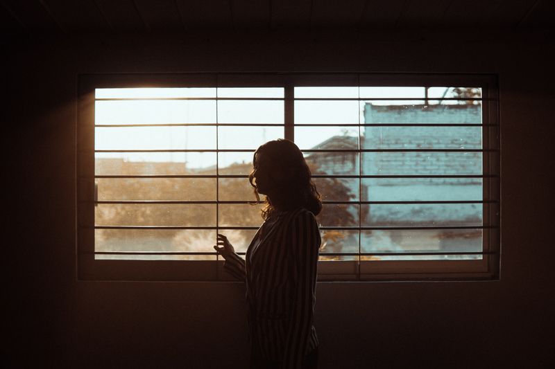 Woman stands by a window, backlit by the afternoon sun with a building and some weeds in view. 