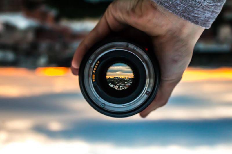 Image: hand holding camera lens focusing in on a city at sunset