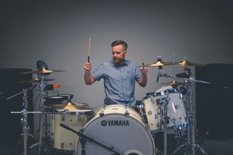 A man with brown hair and a blue button down shirt sitting behind a white drum set with his sticks in the air.