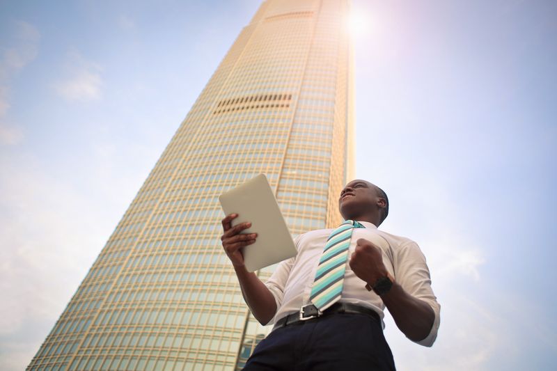 A young man shot from below, wearing a tie, holding a tablet, doing a fist pump, in front of a tall building.