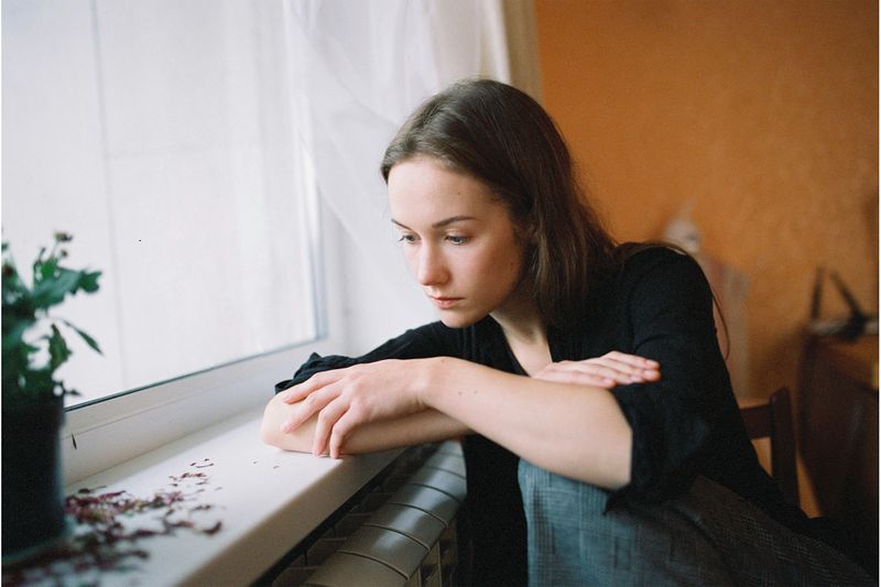 A young woman thinking beside a window in a bedroom.