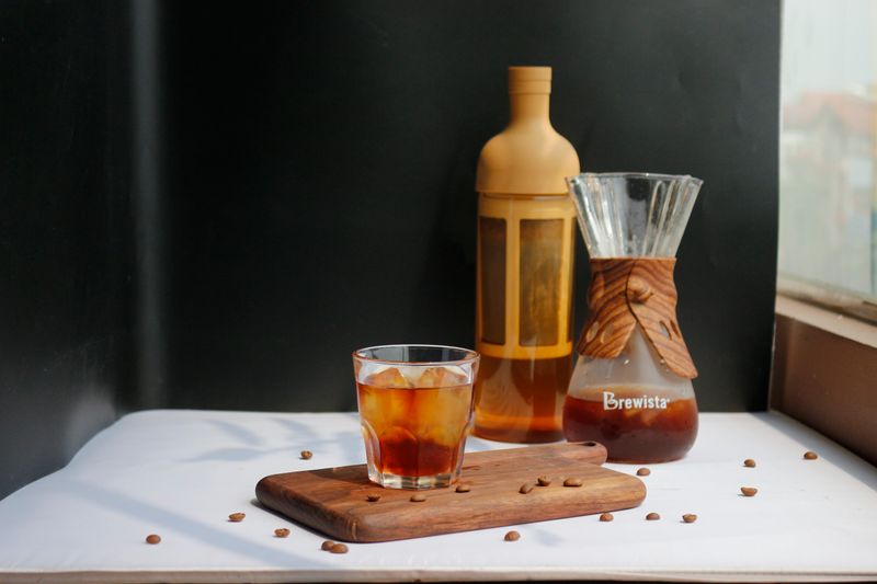 an image of a cup of cold coffee on a cutting board with beans and cold brew press in the back.