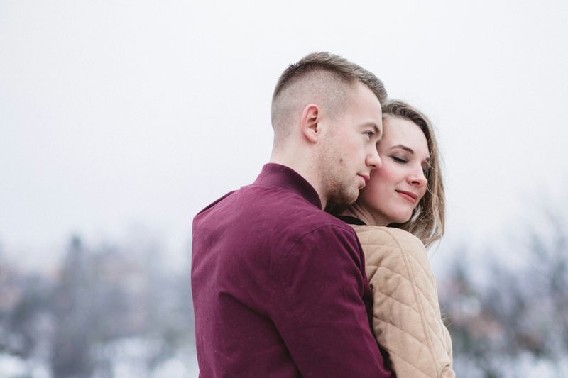 A man in a maroon shirt hugging a woman in a beige top from behind, both with calm smiles on their faces.