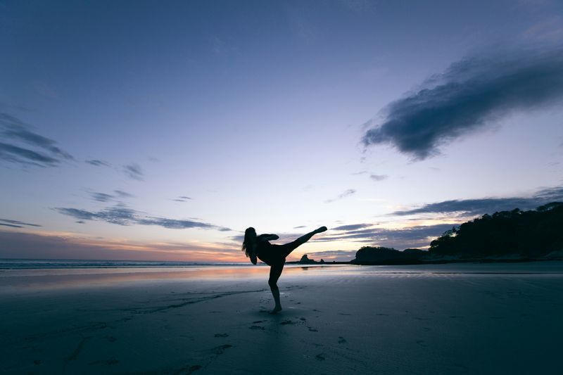 A woman in silhouette on a beach doing a high kick.