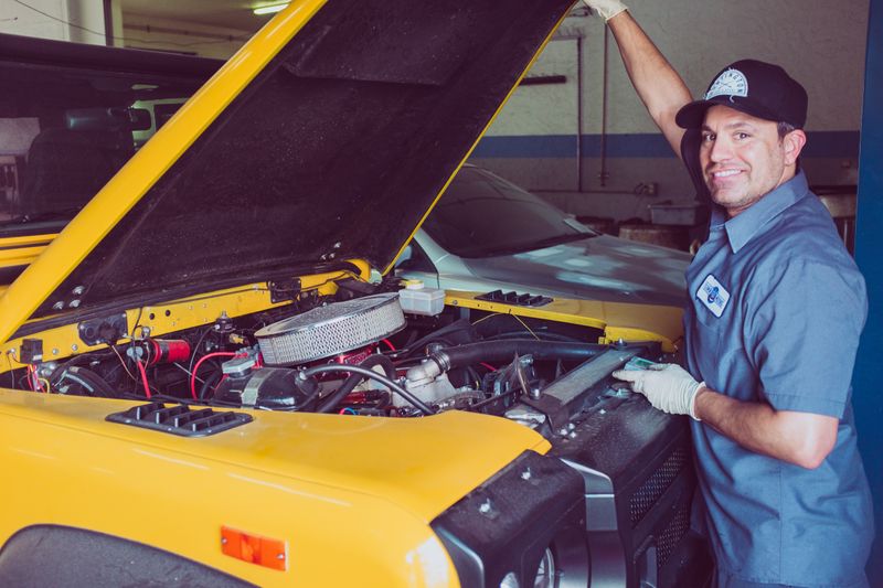 Man in a blue shirt standing in front of a yellow car with the hood up looking at the camera and smiling.