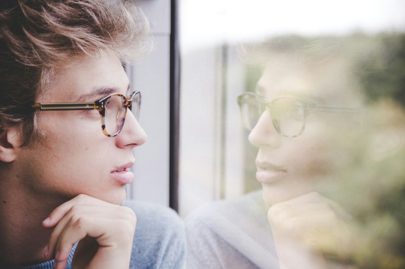 A young man with glasses staring out of a window, showing his reflection.