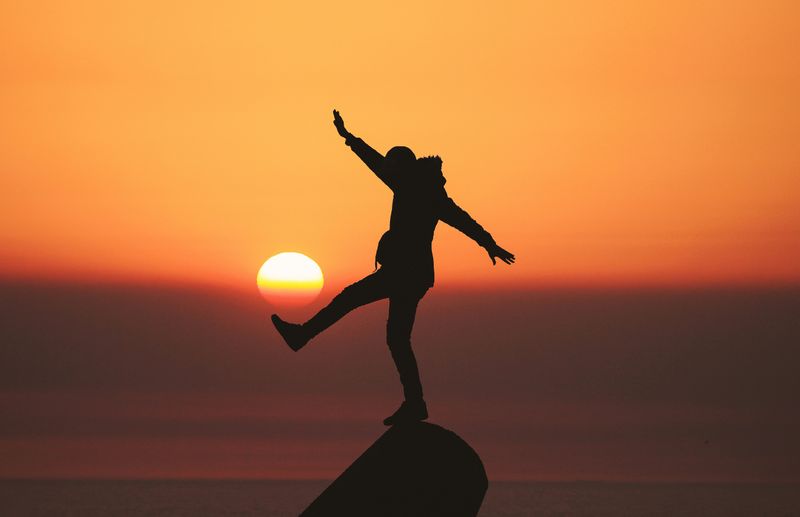 A silhouette of a person balancing on a rock against the sunset.