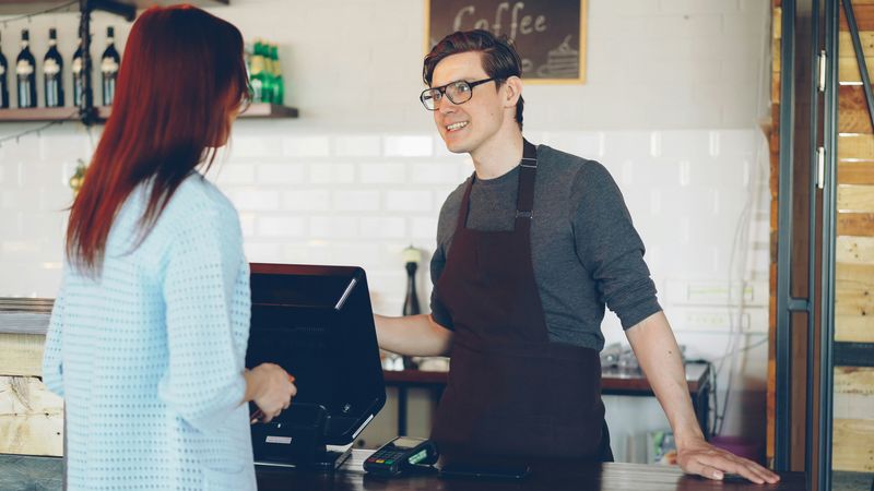 A man stands behind the counter in a coffee shop, talking to a customer.