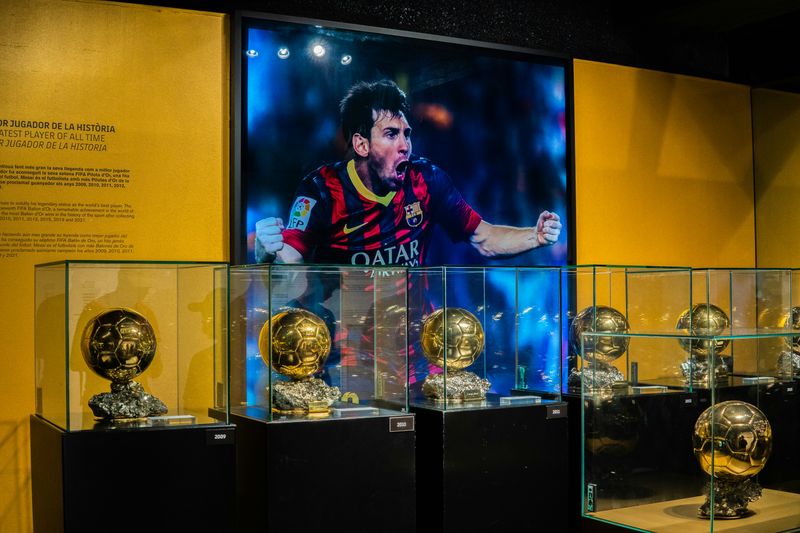 A sports museum room with a large image of Lionel Messi on the wall. Several trophies are in glass displays.