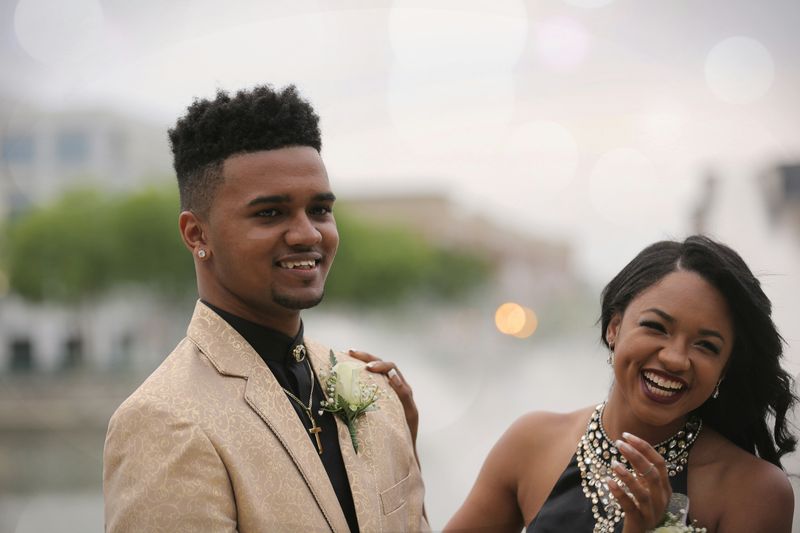 A happy couple dressed in stylish clothes. They both wear corsages and pose in front of an outdoor scene.