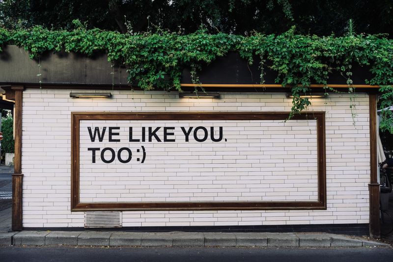photo of a white brick building exterior with a brown frame around the words we like you too with smile emoji 