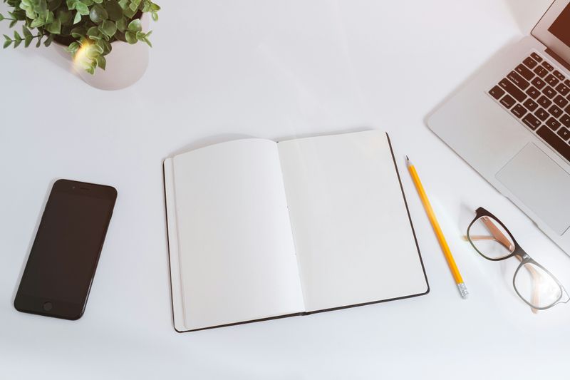 A notebook on a desk with a pencil next to it, as well as a phone, glasses, and a laptop.