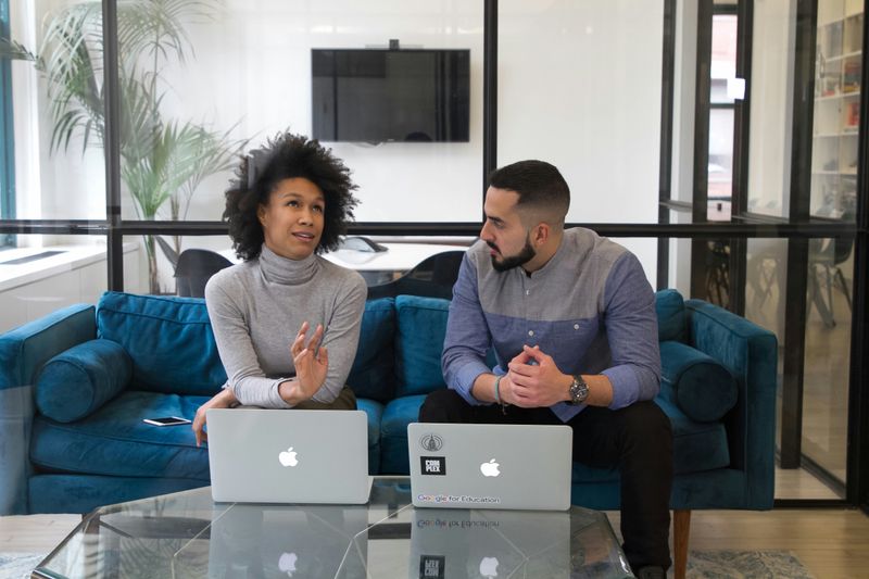 A woman and a man at work, sitting on a sofa with laptops on the table in front of them, having a conversation.