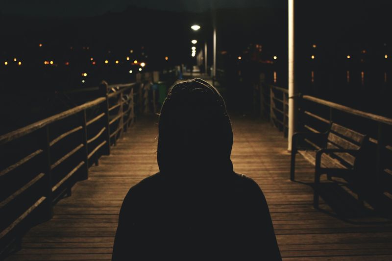 Person wearing hoodie looking down an empty, not well-lit wooden pier at night