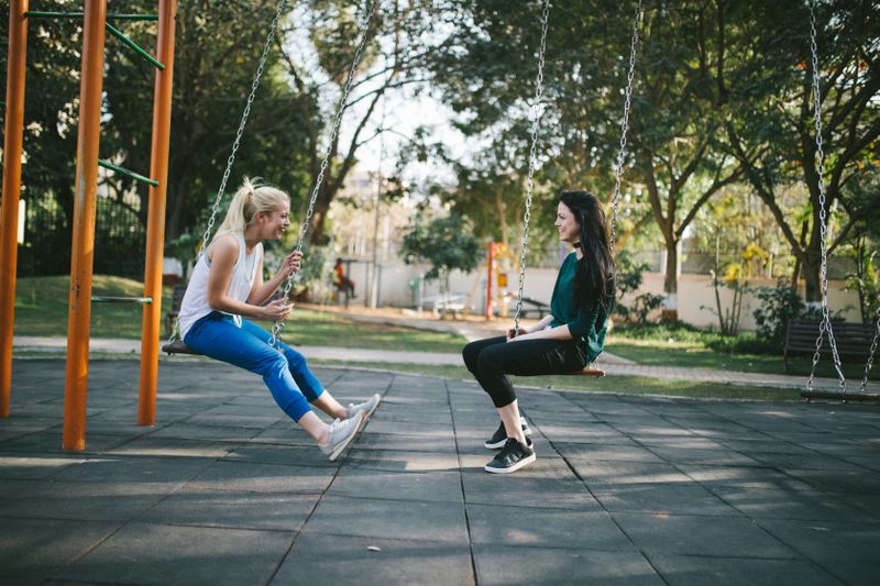 Two young women sitting on swings while talking to each other in a park.