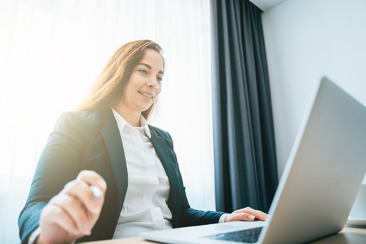 A woman in formal attire in front of a laptop in a bright room