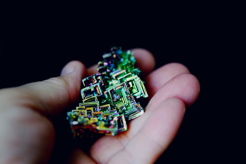 A hand holding a sample of bismuth. The bismuth has cube-shaped crystals with a rainbow-colored film on the outside. 