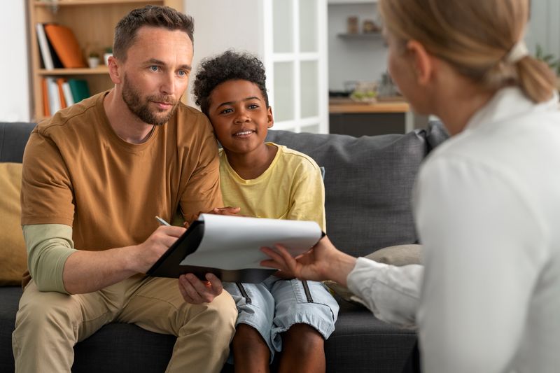An adult and a child sit together on a couch while another adult holds a notepad, discussing ideas.