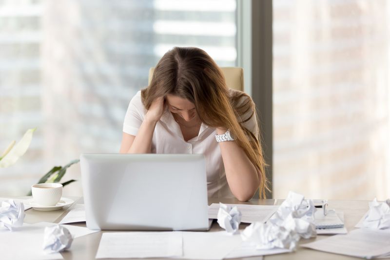A stressed woman in front of her laptop. Piles of scrunched paper litter her desk.