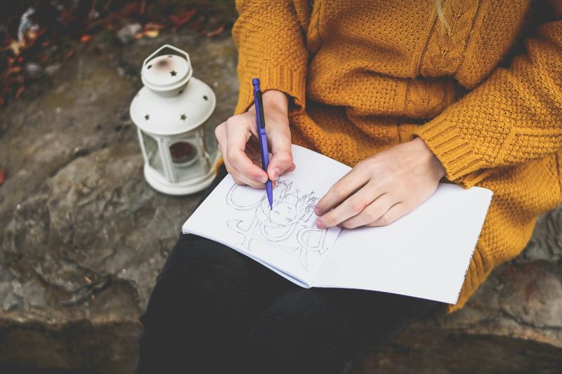 A woman drawing in a notebook with a pencil while sitting on a rock outdoors.