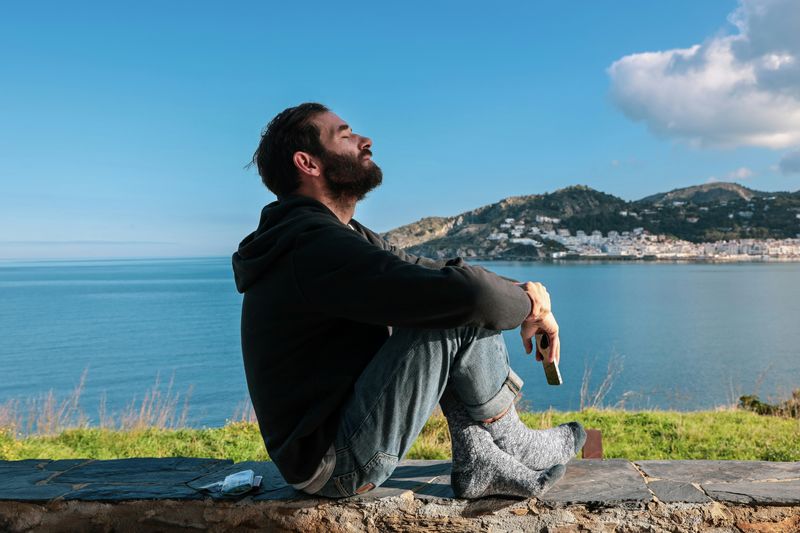 A man calmly sitting on a stone ledge beside the sea with his eyes closed. 