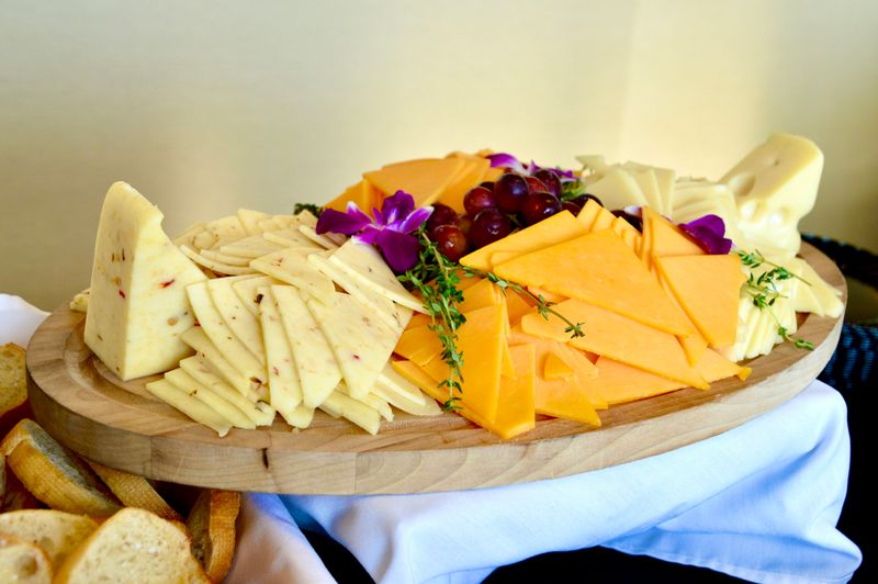 A cheese board with pepper jack, cheddar, and Swiss. There are grapes and flowers in the middle and bread off to the side.
