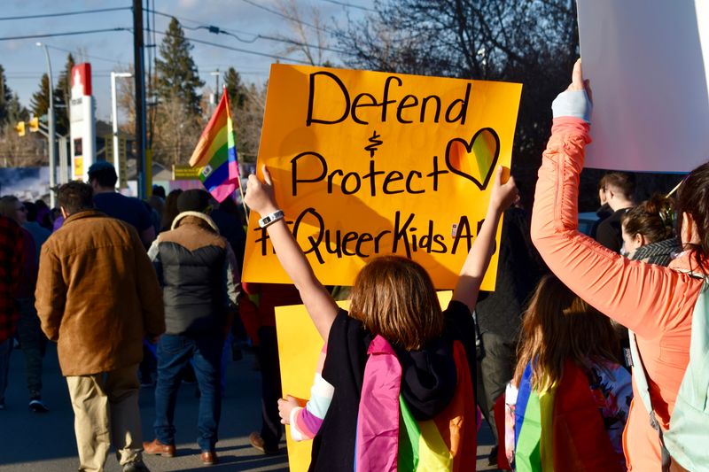 A young person at a rally holds sign saying 'Defend & Protect Queer Kids'.