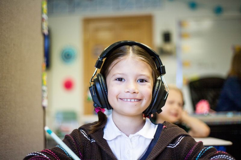 A smiling girl listens on headphones while her classmates and teacher continue to work behind her.