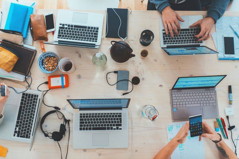 Coworkers collaborating at a desk with laptops, mobile phones, and headphones.