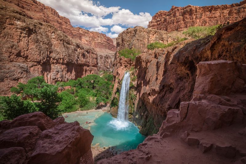 A waterfall with clear blue water, surrounded by trees and canyon walls made of red rock.