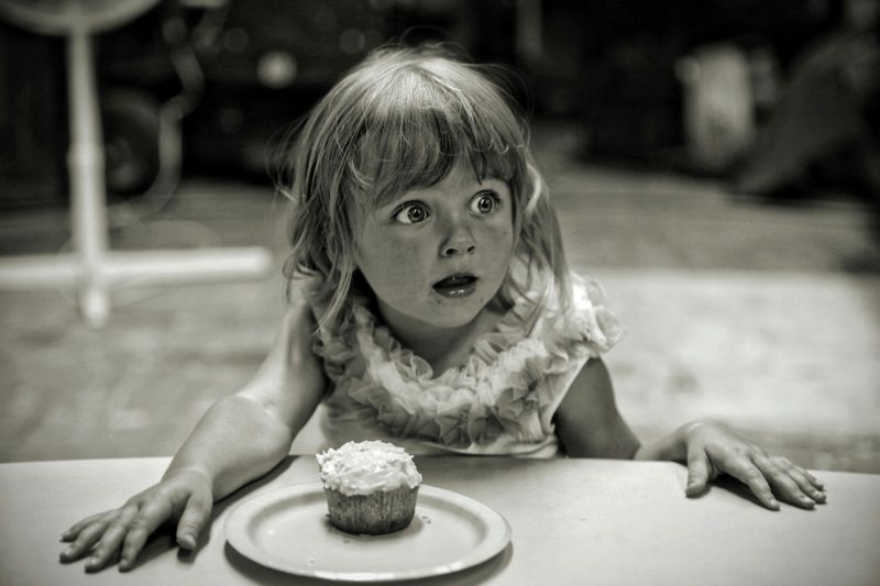 A young girl with astonished expression with a cupcake on table in front of her as she looks at someone offscreen.