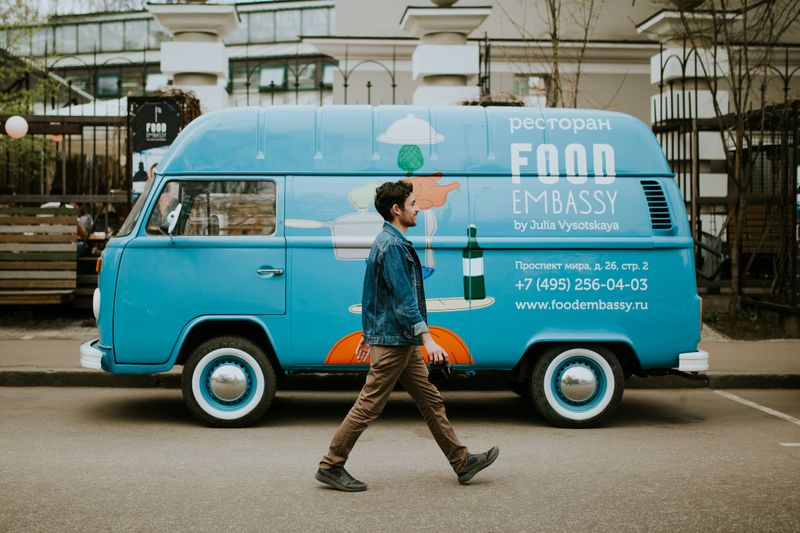Young man in denim jacket, brown pants, and black shoes walking in front of a blue van that says food embassy on the side