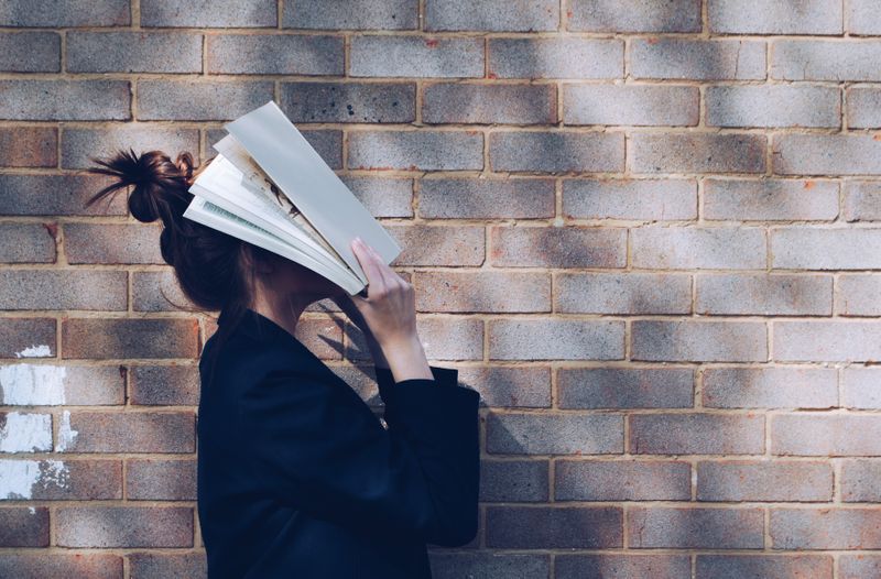 a woman hiding her face with a book, standing against a brick background