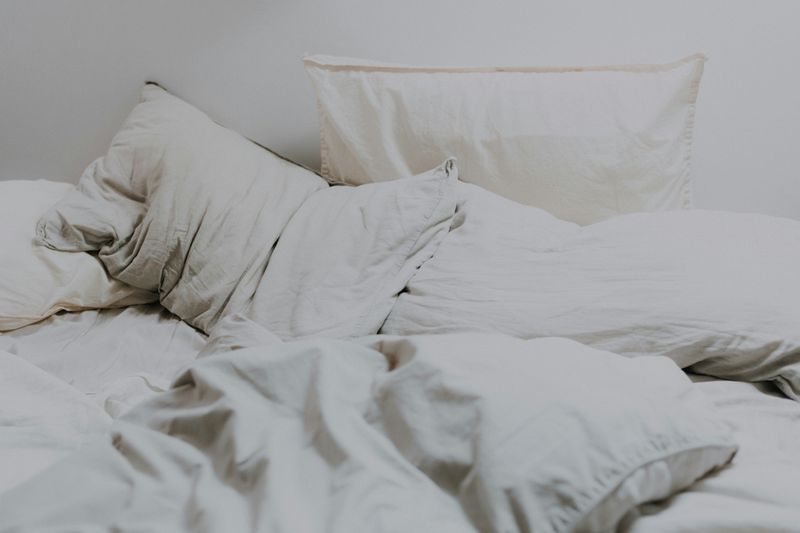 A bed with white sheets, a thick duvet, and about 4 pillows messily made with a white background.