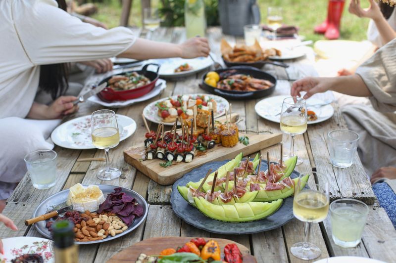 A picnic table with glassware, ceramic serving dishes, and metal utensils all full of fresh food.