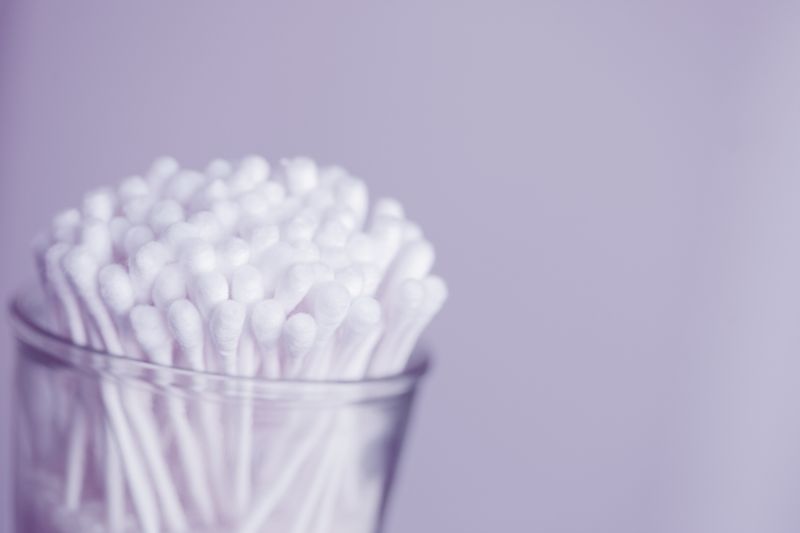 A clear canister full of new cotton swabs sits in front of a light gray background. 
