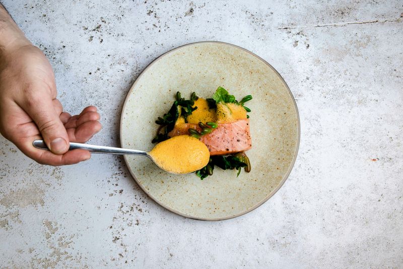 A person using a spoon to put sauce onto a plate with roasted salmon and leafy greens. 