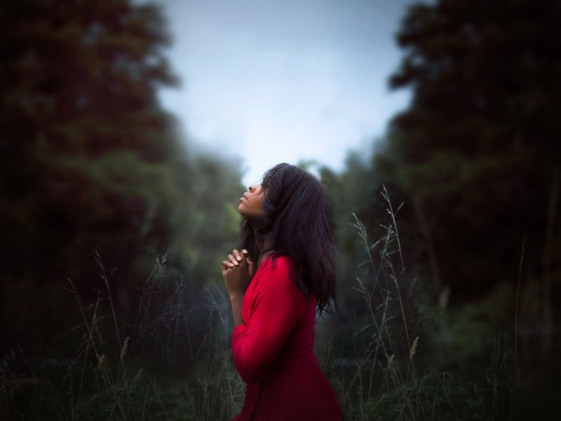 A woman with a pleading face closes eyes and clasps palms as she looks up to the sky with a dark outdoor background. 