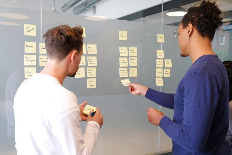 An operations manager helping a colleague arrange ideas on a board with sticky notes.