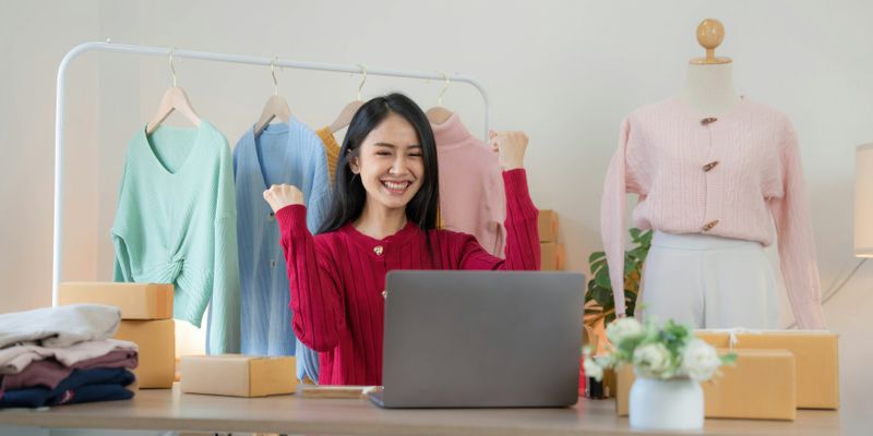 A woman in her clothing shop smiling at a computer. She considers using AI for logo design.