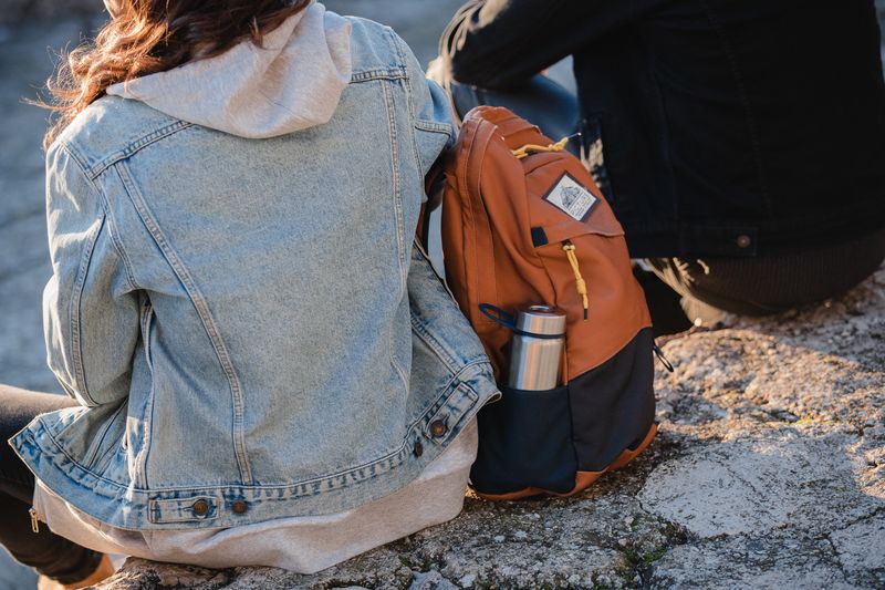A student sitting outside with an orange knapsack with a water bottle.