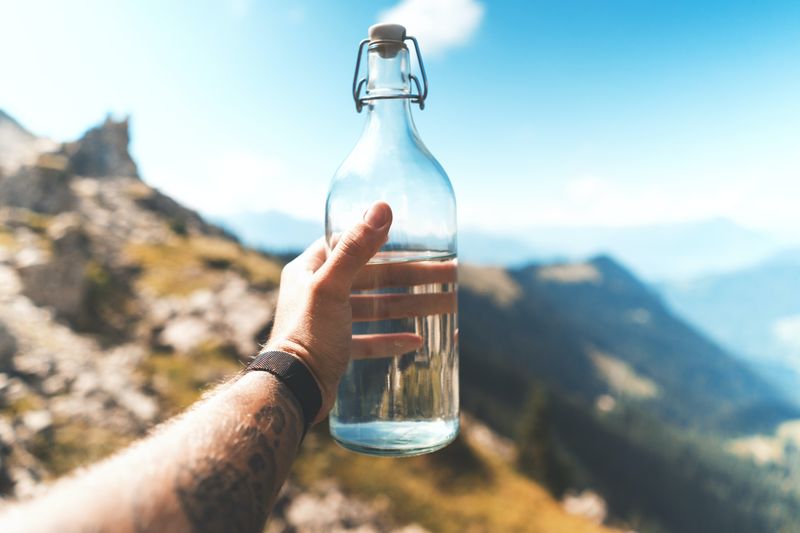 Image of person holding a glass bottle containing water with mountainous background.