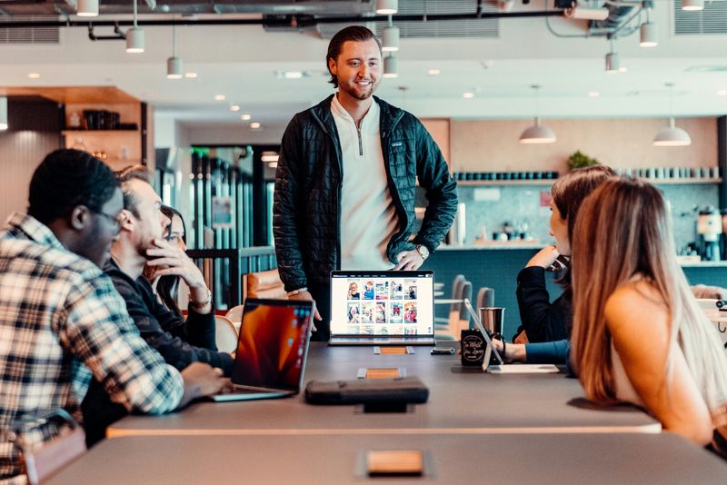 A diverse team meeting around table, with a smiling leader standing at the end of the table with an open laptop.