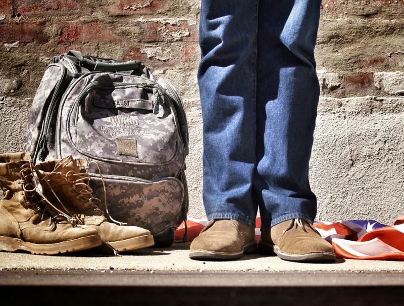 A person standing among military equipment: combat boots, a camo backpack, and an American flag.