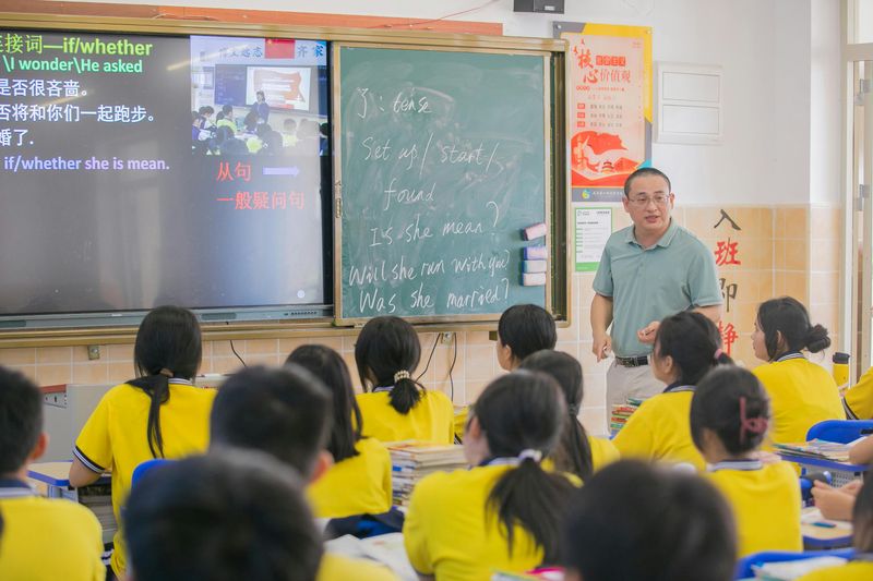 A teacher standing in front of a class teaching English. There's a blackboard with English phrases and Chinese characters.