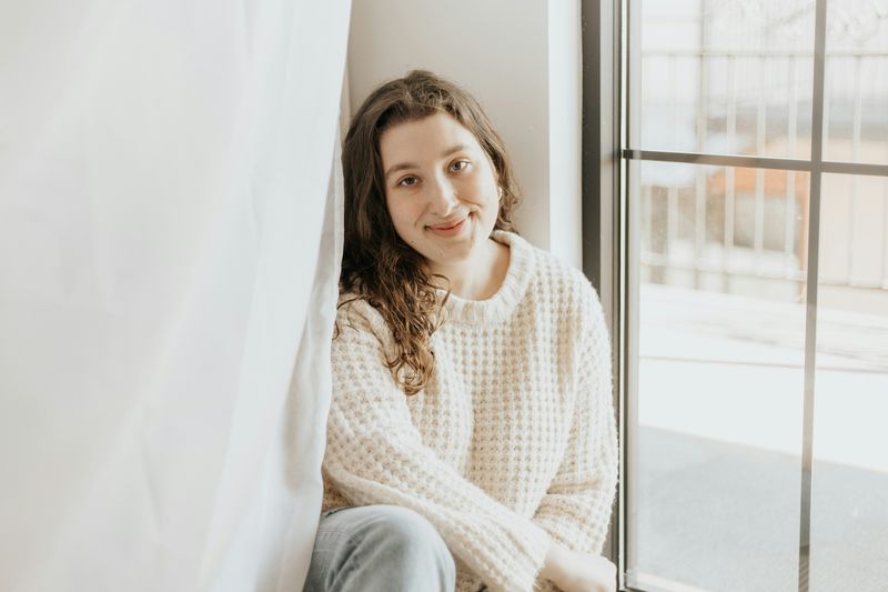 A young woman with log brown hair wearing casual clothing and sitting by a window, looking at the camera.