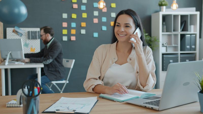  A woman sits in an open office, smiling as she talks on the phone.